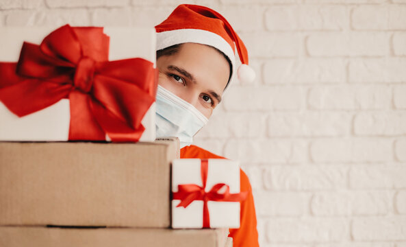 Happy Delivery Man In Santa Hat, Medical Face Mask, Protective Gloves Look From Behind Cardboard Gift Boxes With Red Bow. Young Funny Festive Man Courier In Face Mask Carry Christmas New Year Presents