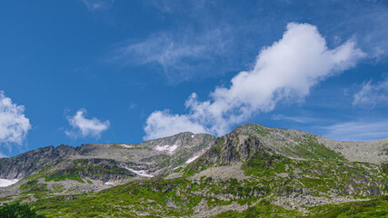 Fototapeta na wymiar Rock ridge with snow and stone placers under blue sky. Summer trip to mountain valley. Atmospheric alpine landscape with stony meadow with green grass.