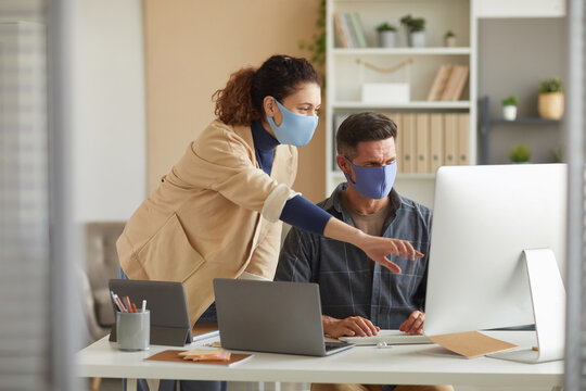 Business colleagues in protective masks working over online presentation on computer in team at office