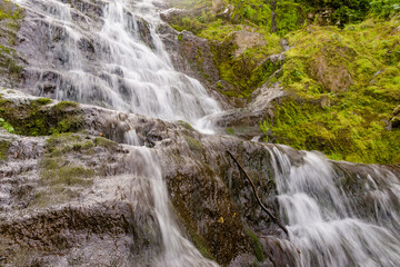 Small cozy waterfall in forest on sunny day. Mountain river falls from cliff onto rocks, rushing stream of water.