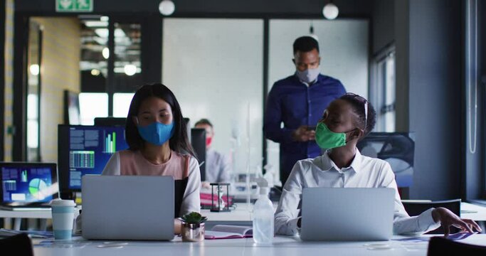 Diverse female colleagues wearing face masks greeting each other by touching elbows at modern office