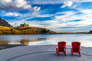 Red chair look over Waterton Lake Marina Point in autumn foliage season sunny morning. Blue sky with colourful clouds reflect on the lake water surface. Waterton Lakes National Park, Alberta, Canada.