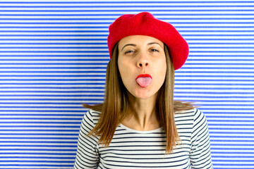 In front of stripped background staying woman with same pattern on t shirt. She is wearing red hat and lolls out to photographer. Facial expression.