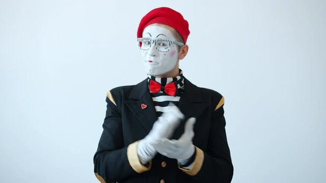 Slow motion of cheerful mime artist in funny colorful costume and make-up clapping hands pointing at camera smiling standing alone on white background