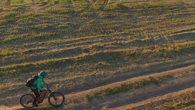 Man in green helmet extreme fast rides on orange mtb bike electro full suspension mountain bicycle e-bike along rural dirty road dust from the wheels past field on sunny autumn evening aerial view.