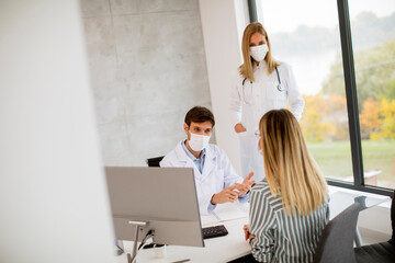 Medical couple doctors with protective medical masks talking with female patient and using computer in the office