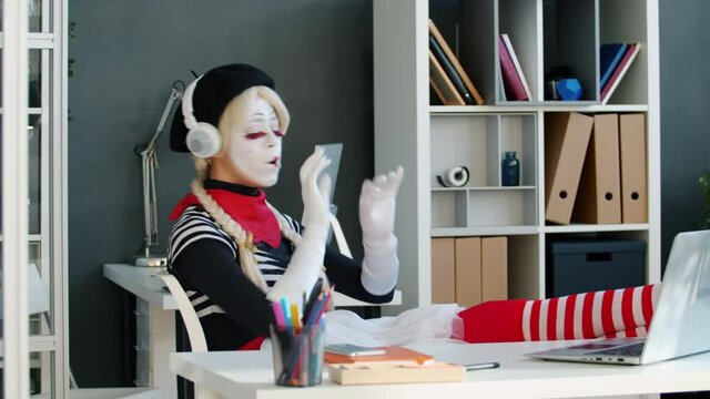 Joyful girl mime in funny costume is enjoying music wearing headphones singing at desk in office. Modern technology and entertainment concept.