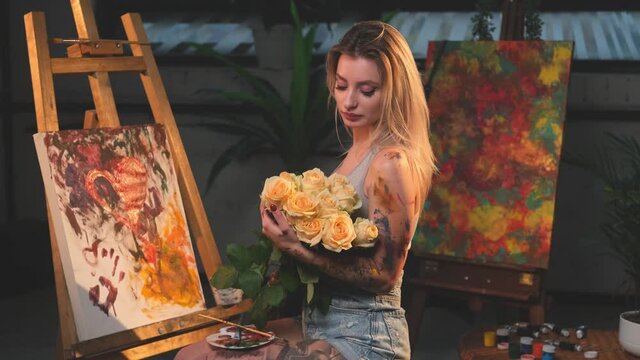 Grimy and seductive female artist with blond hairs poses in dark and spacious studio room holding bouquet of roses and sitting on chair around canvas and artwork.