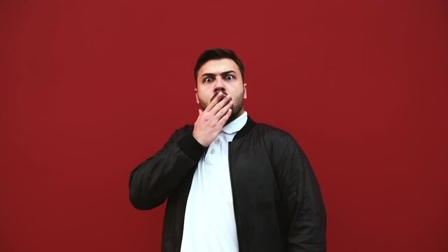 Portrait of a shocked bearded man in casual clothes on a background of a red wall, looking at the camera and surprisingly covers his mouth with his hand.