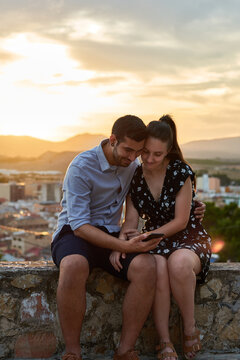 Gentle couple sitting on stone border and embracing while watching video together on cellphone on background of old city under sunset sky