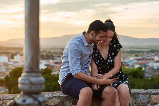 Gentle couple sitting on stone border and embracing while watching video together on cellphone on background of old city under sunset sky