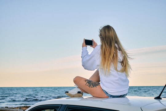 Back view of unrecognizable female tourist sitting on roof of car with smartphone and taking photo of amazing sunset over calm sea - Powered by Adobe