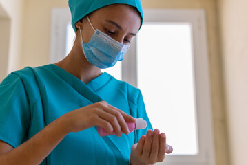 Low angle of young nurse in blue scrubs and medical mask applying sanitizer on hands while preparing for procedure in clinic