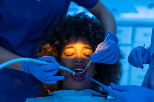 Crop of dentist and assistant using dental suction device on teeth of black woman with closed eyes in clinic