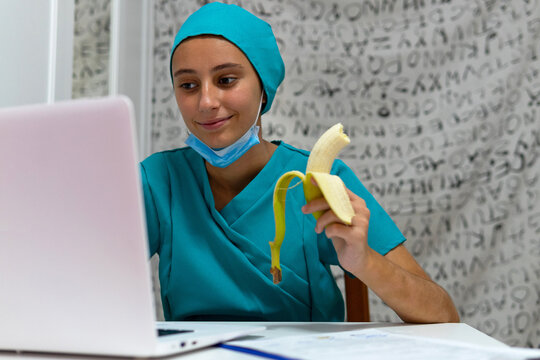 Positive young female medical worker in blue uniform using laptop and eating banana while working at nurses station in hospital