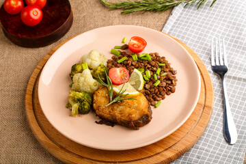 Baked fish with broccoli and lentils in a beige plate on a round stand next to tomatoes and lemon.