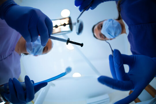 Blurred female dentist with mirror and drill and assistant with suction device standing against clinic ceiling during curing procedure
