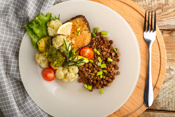 Baked fish with broccoli and lentils in a white plate with rosemary and lemon on a round stand next to a fork.