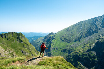 Back view of distant unrecognizable male hiker with backpack and trekking stick standing on grassy hilltop and admiring spectacular scenery of mountain ridge in summer day