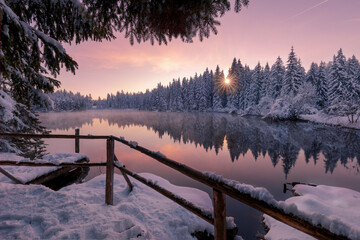 Scenic winter landscape of calm lake with wooden pier covered with snow and coniferous forest reflected in water in sunset time
