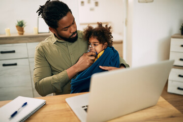 Black father using nebulizer while giving his daughter inhaling therapy at home.