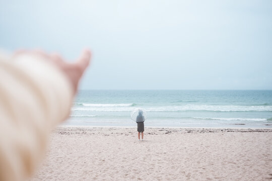 Crop anonymous person pointing finger away towards stormy ocean with lonely traveler with umbrella standing on sandy coast in overcast weather