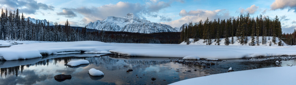 Picturesque panoramic scenery of tranquil river flowing through snowy valley with spruce trees against rocky mountains and blue cloudy sky in wintertime