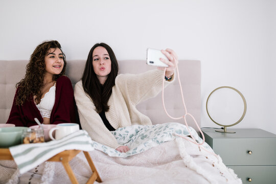 Two friends in bed taking a selfie with the mobile phone. They have dark hair and are wearing a cardigan and lingerie