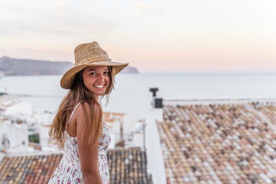 Side view of cheerful female tourist in summer dress and straw hat standing on background of old city near sea and looking at camera at sunset