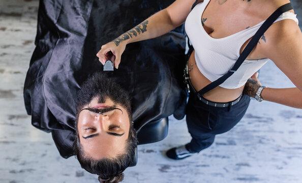 From above of crop unrecognizable female barber with trimmer in tattooed hand shaving beard of stylish brutal client with mustaches in barbershop