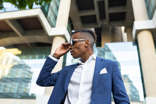 Low angle of masculine African American male entrepreneur wearing elegant suit and sunglasses standing in city and looking away