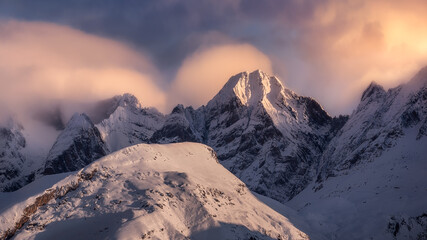 Sharp mountain peaks covered partially with snow and surrounded by fog under picturesque cloudy red sky at nightfall in wintertime