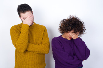 Young couple wearing knitted sweater standing against white wall making facepalm gesture while...