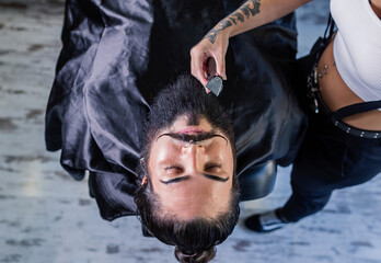 From above of crop unrecognizable female barber with trimmer in tattooed hand shaving beard of stylish brutal client with mustaches in barbershop
