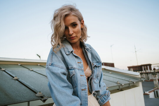 Tranquil female in denim jacket and with wavy hair relaxing in house roof in the city in evening and looking at camera