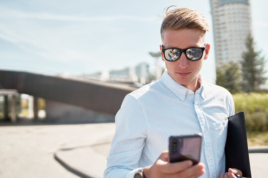 Confident young male in casual shirt and trendy sunglasses carrying paper folder walking on urban street and using mobile phone