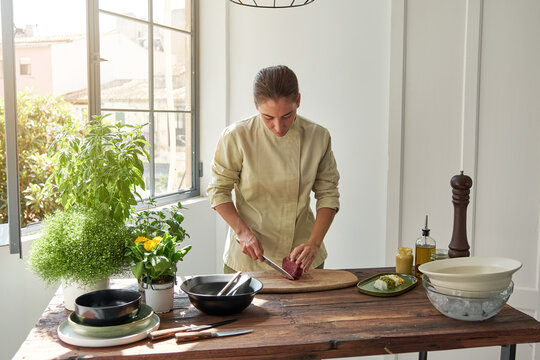 Concentrated female chef standing at table at home and cutting raw beef while preparing Steak tartare