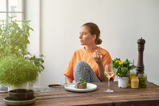 Pensive female sitting at table with Steak tartare on plate and drinking white wine while relaxing at weekend at home and looking at camera