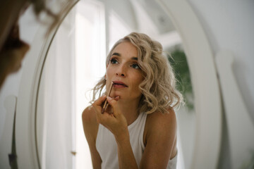 Low angle of charming female applying lip pencil and doing makeup at home while reflecting in mirror