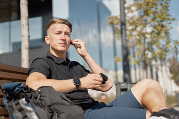 Confident young man in trendy casual outfit sitting on bench near backpack and putting on earbuds while resting on city street