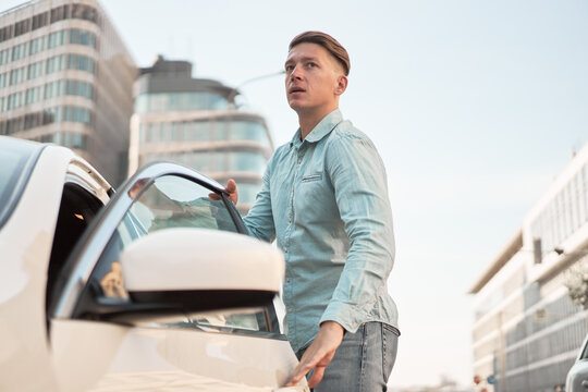Handsome male driver looking away getting in modern automobile while standing on parking in city