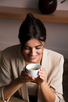 From above of positive dreamy young female with cup of aromatic coffee sitting at table and looking away while relaxing at home