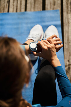 From above back view of unrecognizable female athlete sitting on mat and checking pulse on fitness tracker while resting after outdoor workout