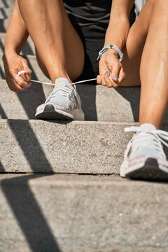 Low angle of crop anonymous female athlete sitting on stone stairs and tying shoelaces on sneakers while preparing for outdoor training