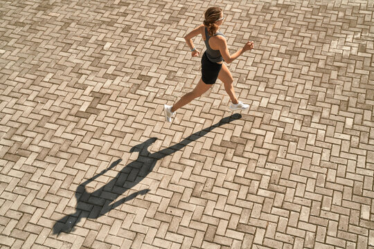 From above full body of active slim female in sportswear running fast on paved square in sunny day