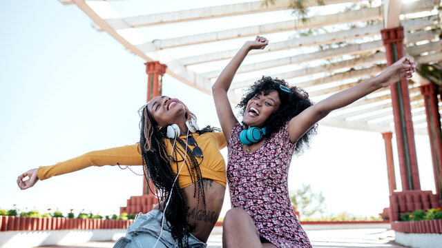 Cheerful black female friends with braids and curly hair sitting on embankment and having fun with raised arms in summer