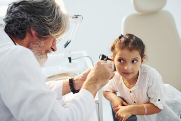 Fototapeta premium Little girl have a visit. Senior otorhinolaryngologist with grey hair and beard in white coat is indoors in clinic
