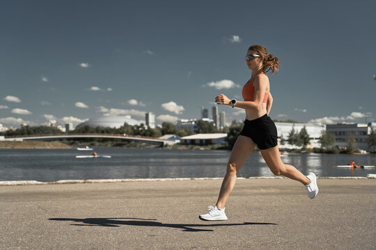 Side view full body of energetic motivated fit female in activewear with fitness tracker jogging on paved embankment near river in city