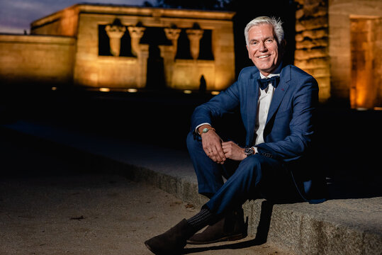 Positive elegant senior male in stylish blue suit with bow tie and wristwatches sitting on stone border in night city and looking at camera