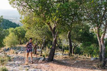 Active young female hiker with trekking poles climbing stony trail on mountain slope with green trees during training in nature